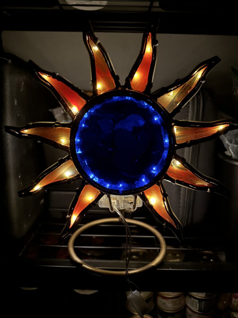 Blue moon-faced center glass surrounded by golden rays holding orange glass. Backlight with fairy lights.