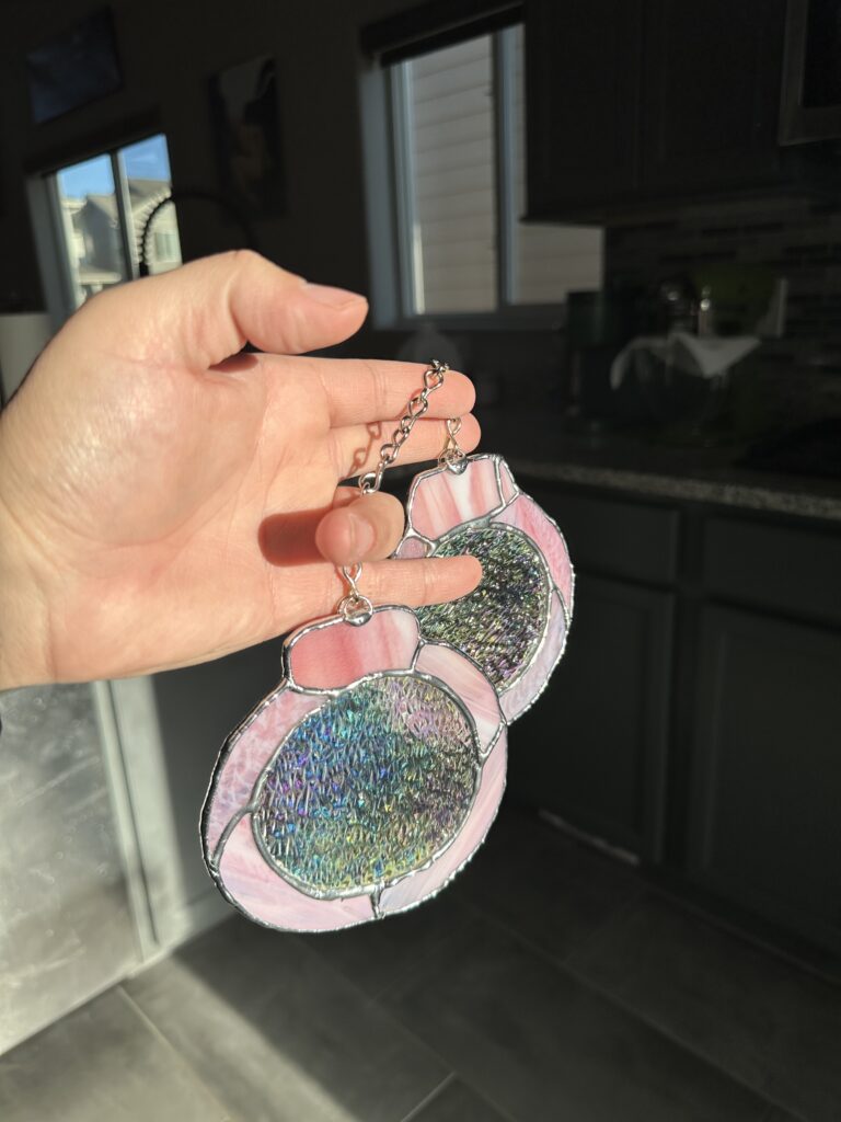 A hand holding up a pair of pink handcuff-shaped glass with iridized clear in the middle of each cuff.