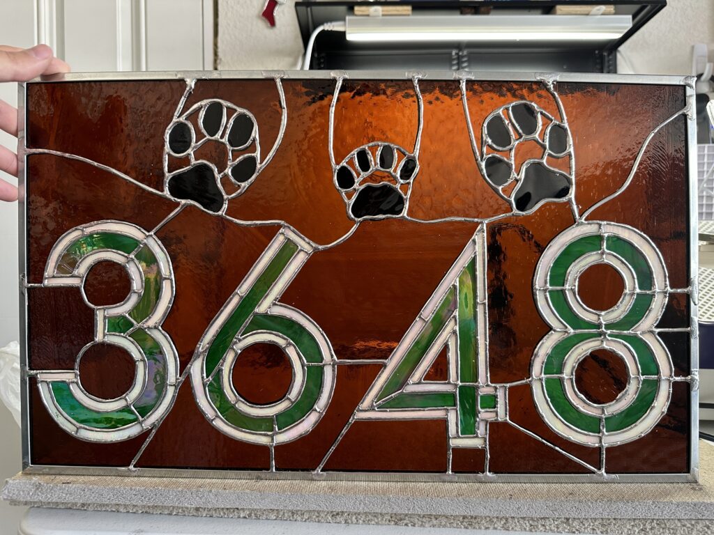 rectangle stained glass panel with iridescent green numbers 3, 6, 4, 8, outlined in iridized white. The background is brown and there are 2 dog and 1 cat paw print above the numbers.