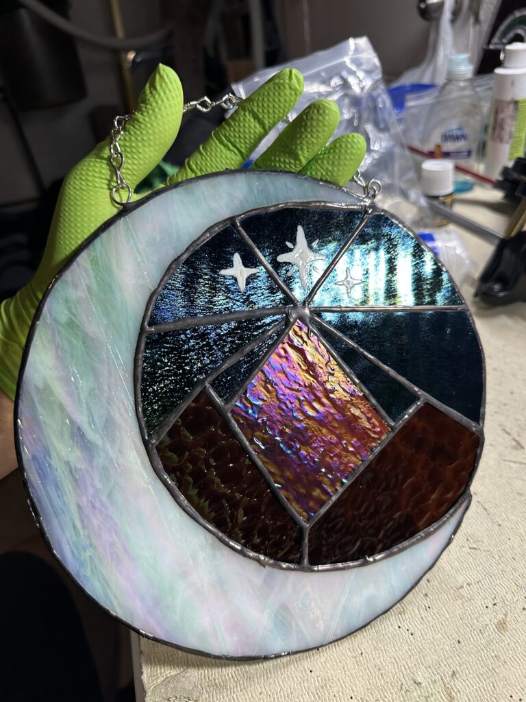 Iridized white crescent moon with 3 mountains sitting inside. There's a night sky painted with 3 stars.