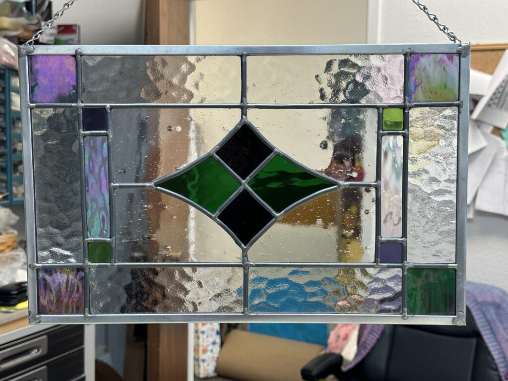 Rectangle stained glass lead panel with a sideways diamond in the center made of purple and green glass. There are green and purple squares in the corners.