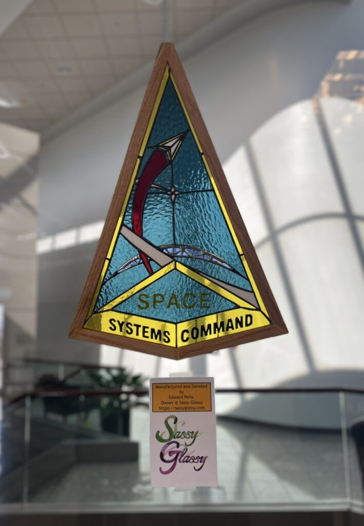 SSC logo hanging in a window. Chevron shape with "SPACE" in yellow paint on blue glass and "SYSTEMS COMMAND" in blue paint on yellow glass. There's a crescent earth with network lines and a small chevron surrounded by yellow glass coming from a red "plume."