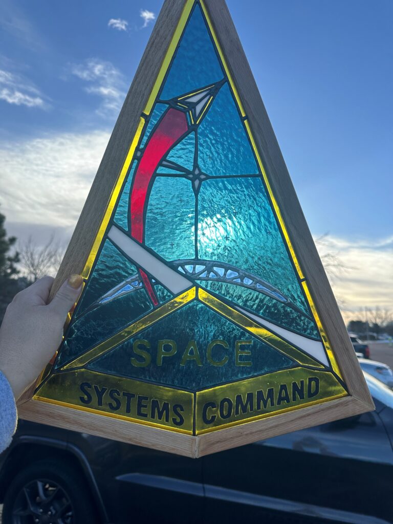 he Space Systems Command chevron-shaped unit logo in glass. Inside the wood frame is a yellow gold border. The dark blue background has small stars painted on and there is a glass 4-pointed star in the center-ish. To its left is a red curved vertical strip of glass leading to a small white chevron outlined in gold. Under it, above the painted “SPACE SYSTEMS COMMAND” words, is a crescent earth with intersecting angled lines. Most of the project except this feature is patina’d black.